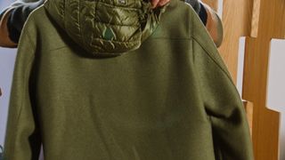 Back image of the green hooded parka style sweater, persons hands adjusting the hood, wood frame to the right, grey background
