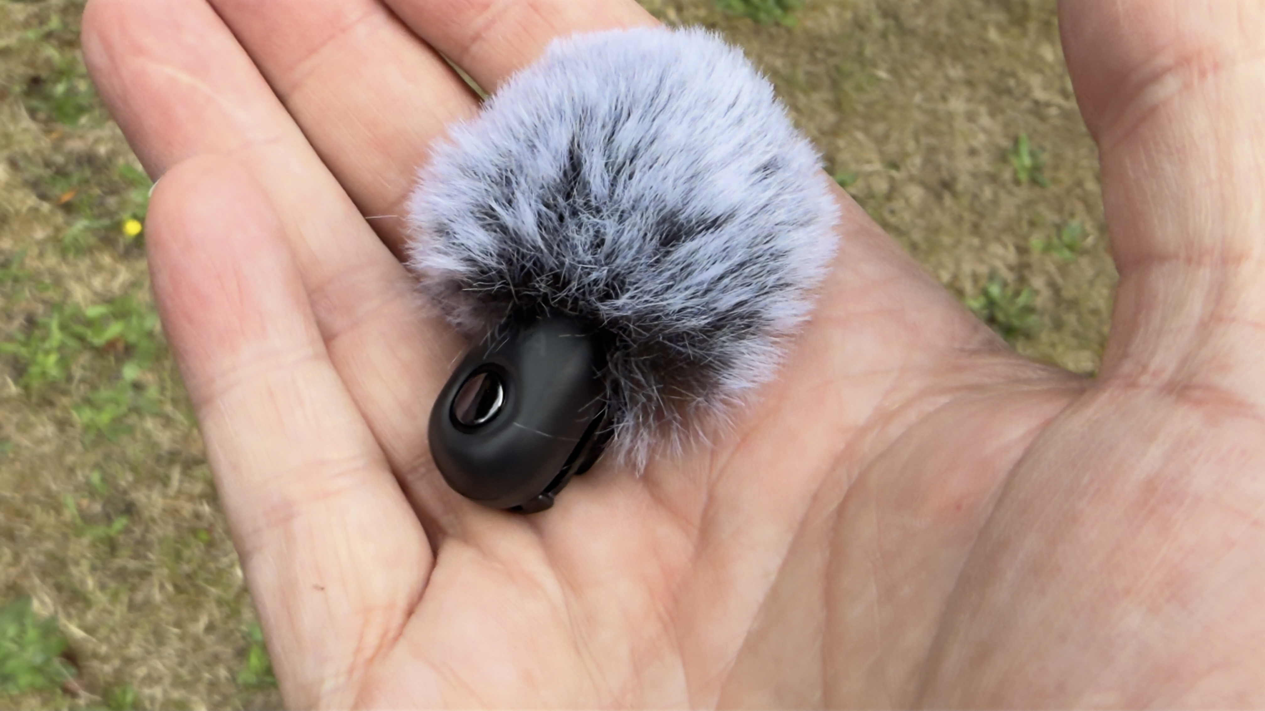 A hand holds one of the two supplied lavalier clip mics with a fluffy wind shield attached