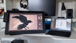 A photo of the Xencelabs pen display set up on a desk with a laptop and a painting on a raven on the screen