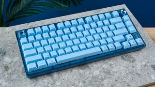 Corsair K65 Plus (Mac) in light blue color, on a stone surface with a blue wall and a green plant in the background.