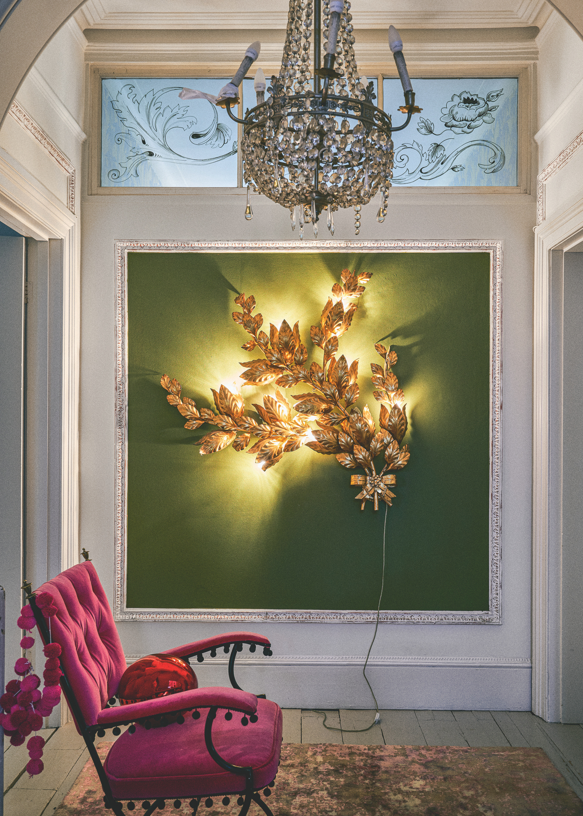 A white hallway with a square feature wall has a gold leaf piece of 3d art lit up with string lights; a pink chair sits sideways in front of the feature with a pink bauble garland draped over the back and a large red bauble decoration sits on the seat