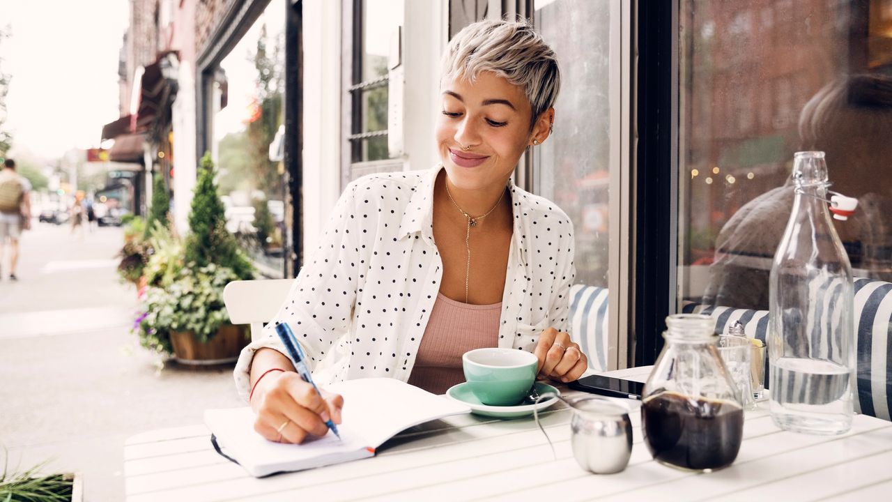 Woman writing in diary at sidewalk cafe to illustrate journaling for beginners