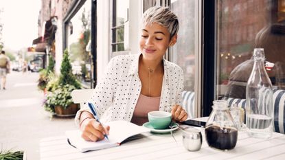 Woman writing in diary at sidewalk cafe to illustrate journaling for beginners