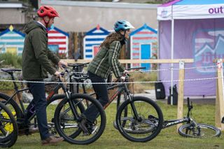 Kate Middleton and Prince William wearing green coats and bike helmets riding bikes on grass with a row of colorful sheds behind them