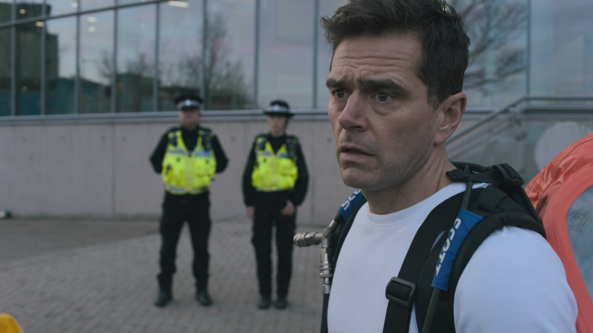 Iain Dean outside the swimming pool looking very worried, wearing a white t-shirt and an orange backpack. Two police officers can be seen standing in the background.