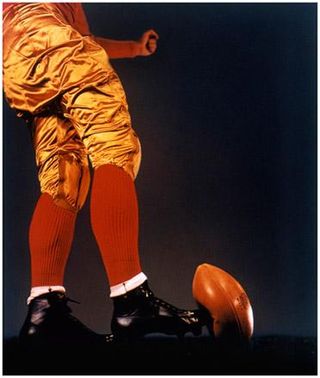 Photograph in gold and orange tones with black background showing the lower half of a footballer in old-fashioned kit wearing plain black boots about to kick the ball.