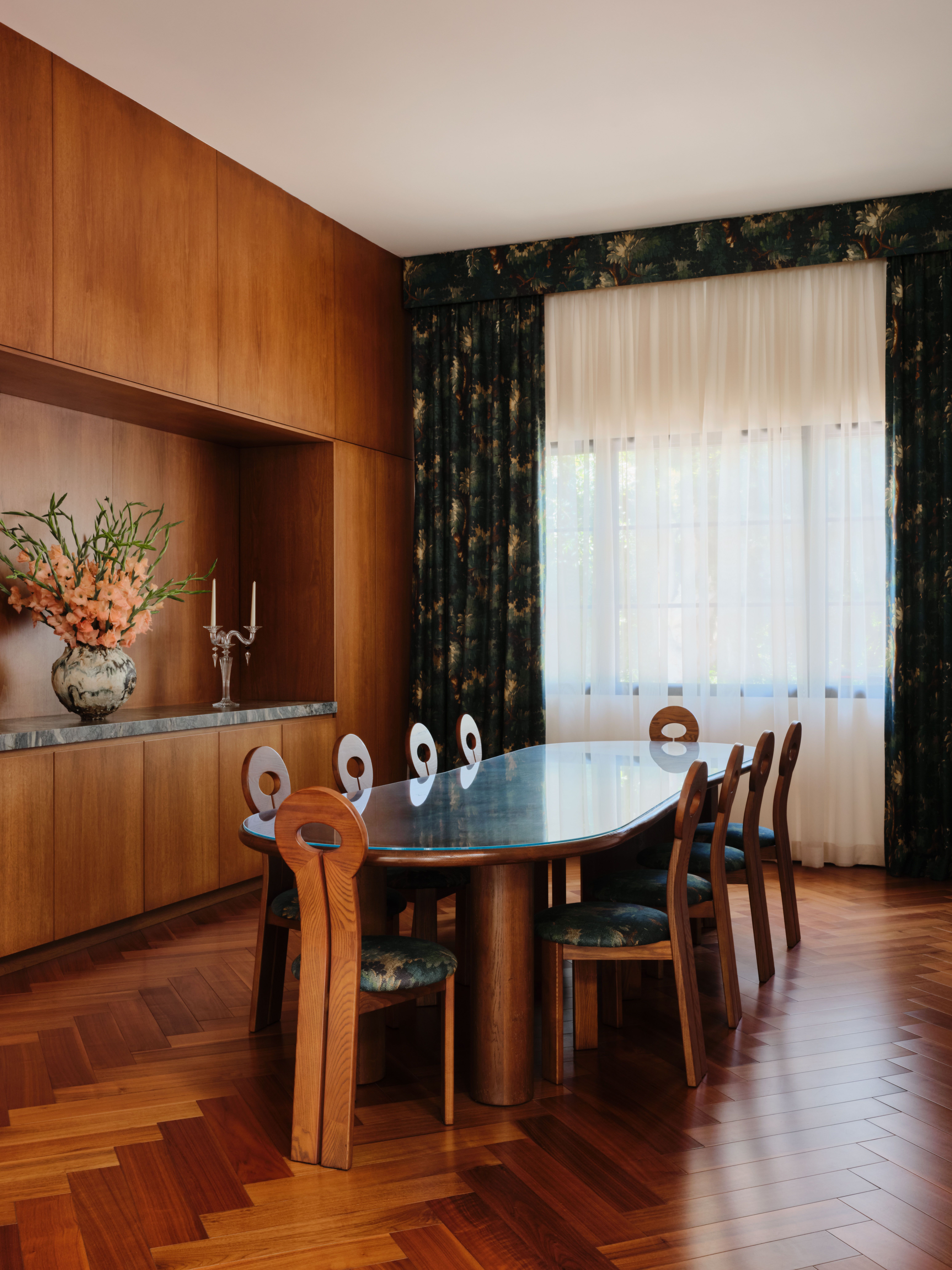 Dining room with herringbone flooring, a wooden table set with sculptural upholstered chairs, patterned blue and gold curtains, and a huge flower vase set on a built-in shelf on the left