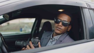 Photo of a man in sunglasses behind the wheel of a car holding a camera