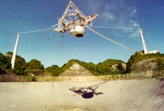 An archive image of Arecibo Observatory's massive radio dish shows the heavy science platform suspended above it, two of the supporting towers and the complex cabling holding it up.