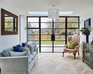 A lounge area with herringbone flooring, a pink armchair and black-framed glass doors