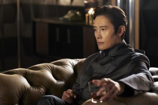 Lee Byung-hun as Front Man, sitting in a large leather chair and holding a glass of liquor, in Squid Game S2.