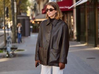 Fashion influencer @monicaainleydlv wearing a chic outfit styled with a brown leather collared coat, cream jeans, leather gloves, and sunglasses.