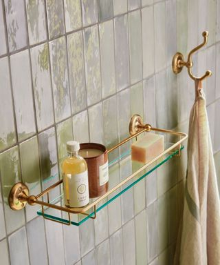 A gold and glass bathroom shelf with candles and toiletries on on pearly tiles