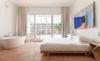 Interior view of the room in the Casa Mãe hotel. Large, airy space, with floor-to-ceiling windows, a large bed with white linen, and a bathtub across from it. 