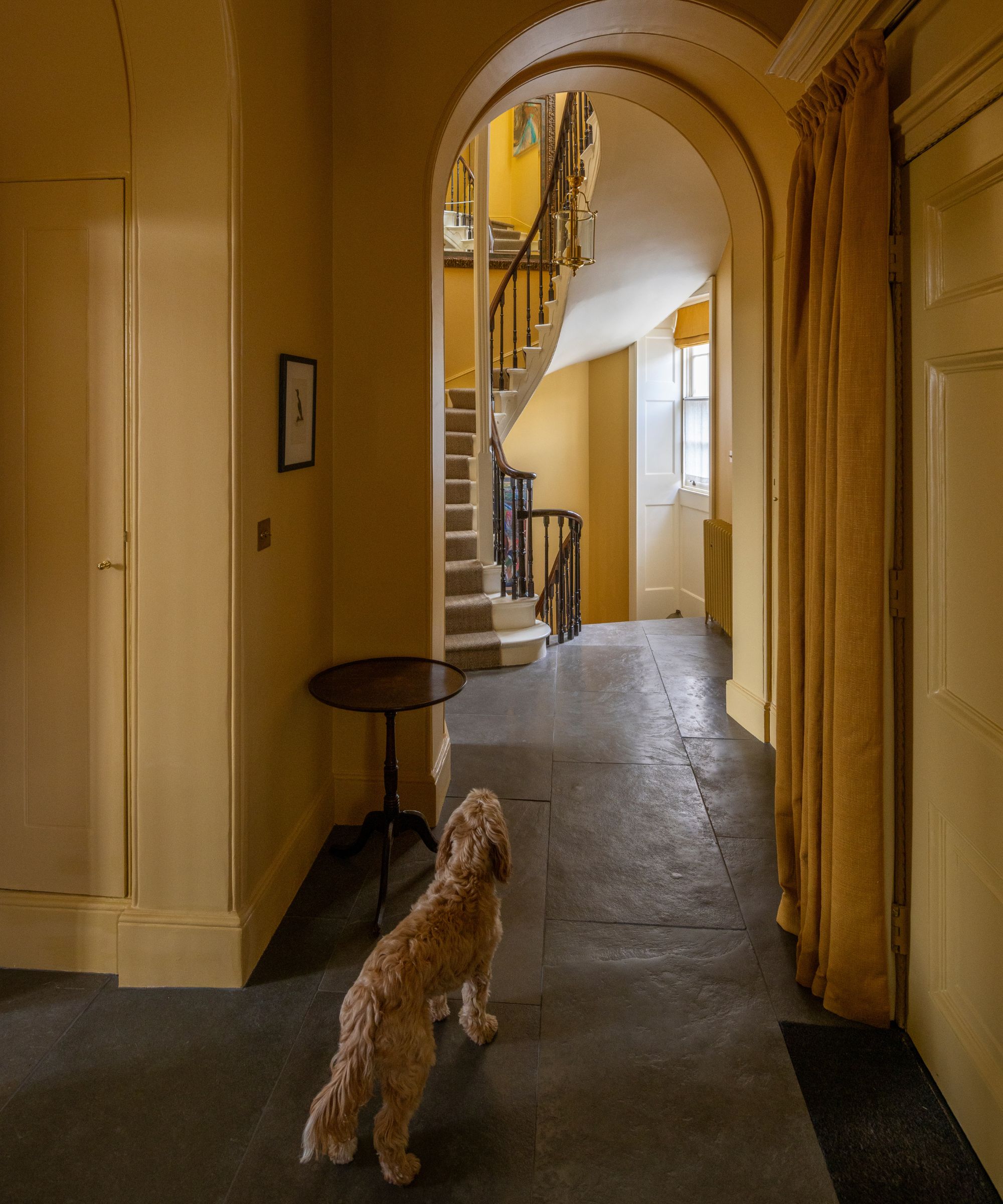 Country house with an arched doorway, walls painted mustard yellow, dark slate floors, and a winding staircase with a wooden bannister and a jute runner.