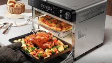 A Typhur sync air fryer oven on a white counter, with a pizza on the top shelf, and someone pulling a roast chicken out on the bottom shelf. 