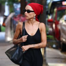 Zoe Kravitz walks down the street wearing a black slip dress with a red bandana, black flip-flops, a black leather bag and oversized earrings.