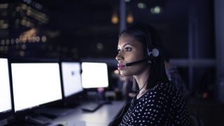 A woman wearing a voip headset while looking at a row of bright computer monitors in a dark room