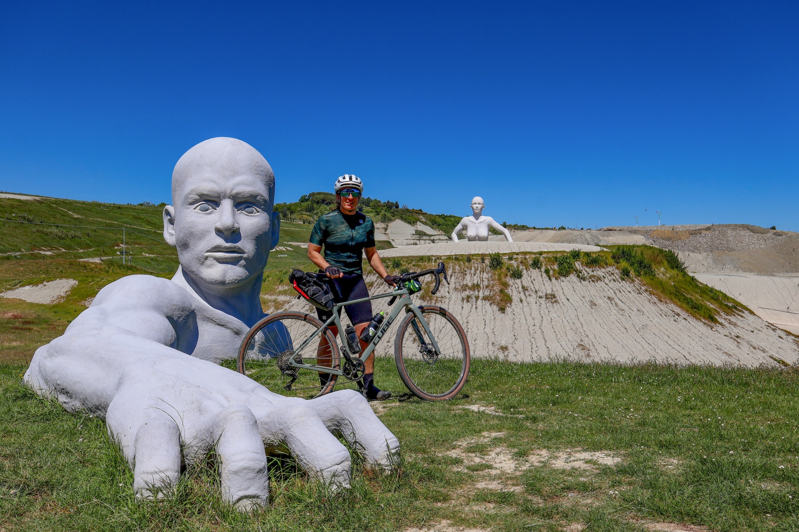 Emma Magnus standing next to a huge sculpture of a man who is emerging from the ground, only his head and shoulders above the ground. He has one arm outstretched, his giant hand in the foreground