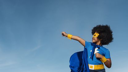 A young black girl dressed in a superhero costume holds out her arms in a gesture of strength