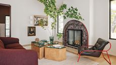 A white living room with wood floor, curved burgundy marble fireplace, burgundy sofa, sculptural burl wood and green glass coffee tables and red and black armchair.