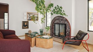 A white living room with wood floor, curved burgundy marble fireplace, burgundy sofa, sculptural burl wood and green glass coffee tables and red and black armchair.