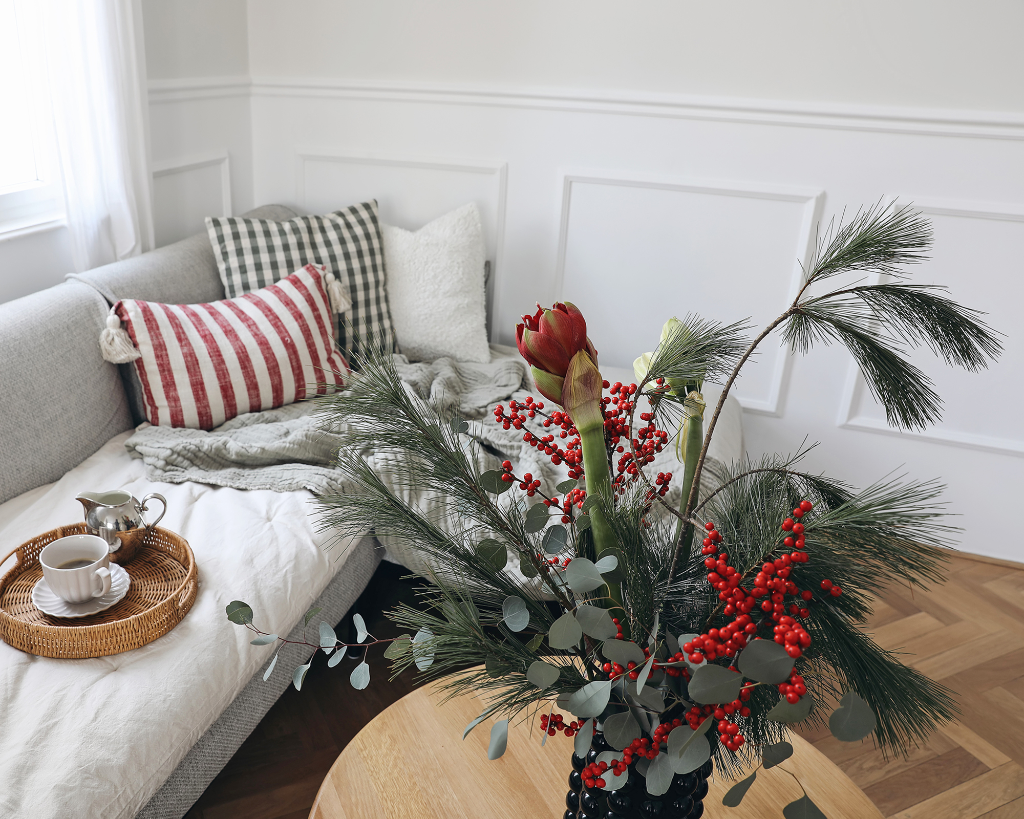 Pine, red holly berry floral arrangement, Blurred background. Scandinavian style sofa with pillows, window, winter morning coffee scene