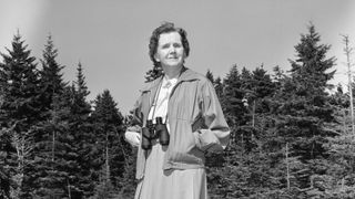 Rachel Carson, American marine biologist and environmentalist, standing in front of some woods with binoculars.