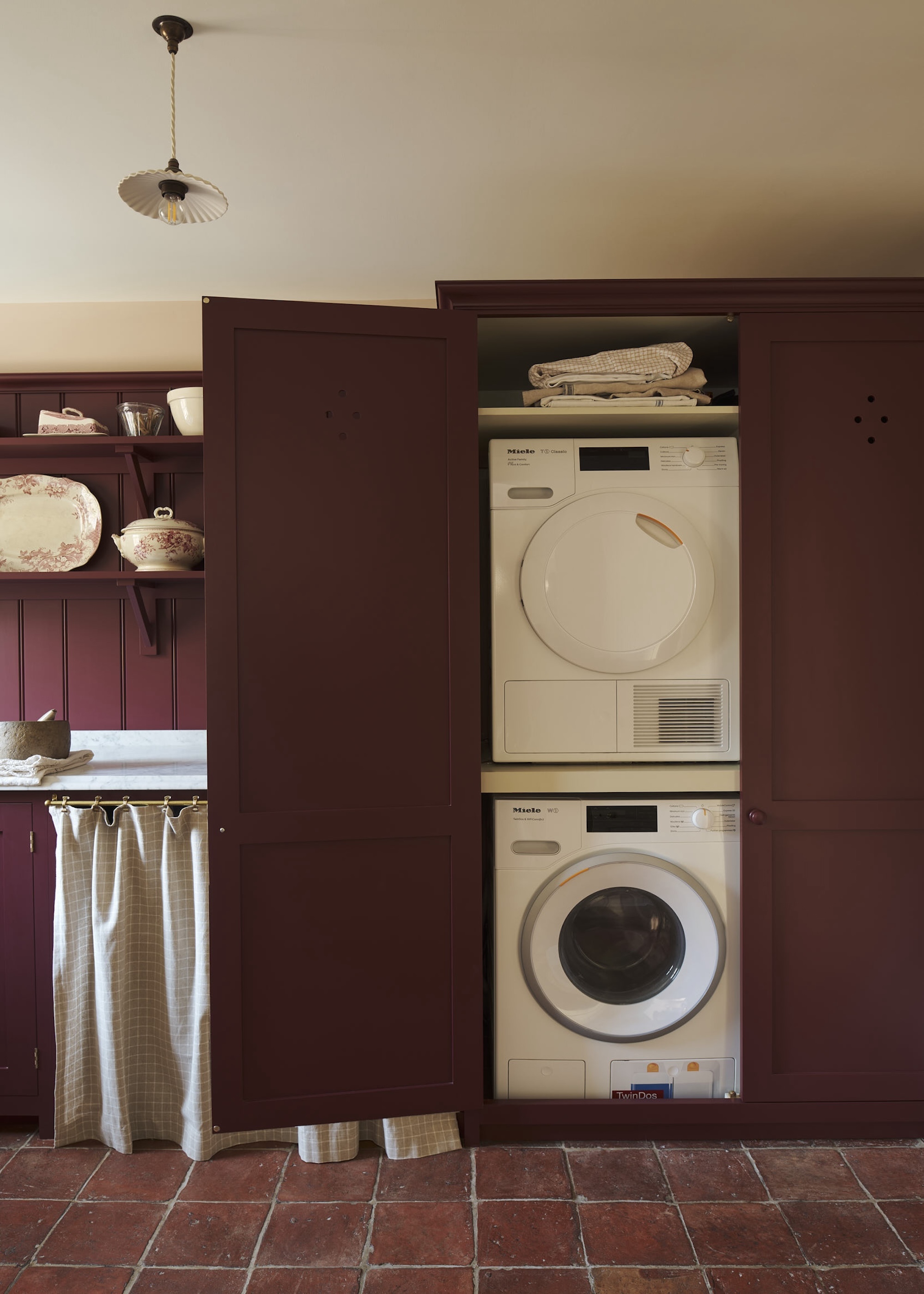 A deep burgundy red utility room with stacked appliances, open shelving and hardwearing tiled floors