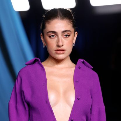 a white woman with dark hair posing on a red carpet. she is wearing a low cut purple dress in front of a blue background