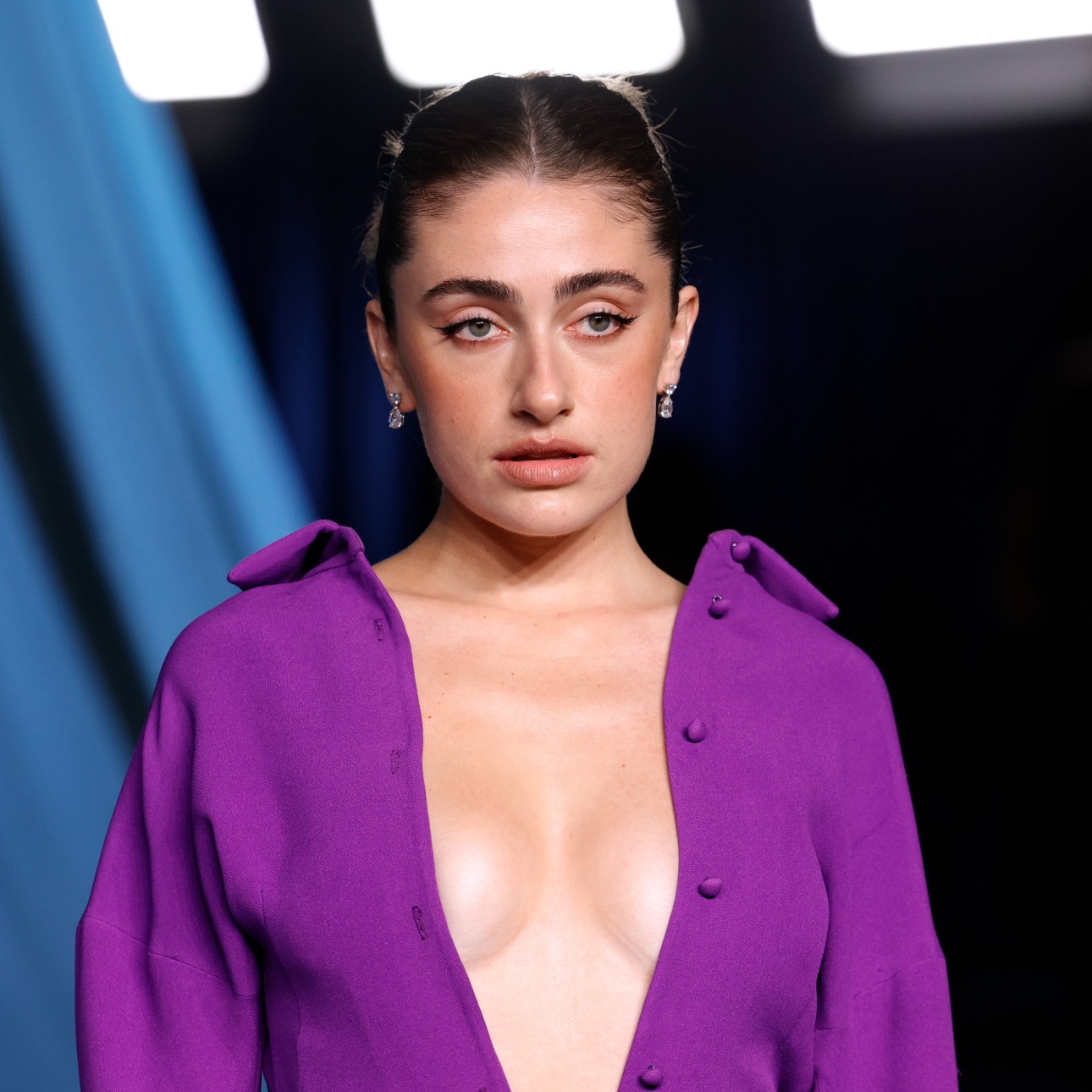 a white woman with dark hair posing on a red carpet. she is wearing a low cut purple dress in front of a blue background