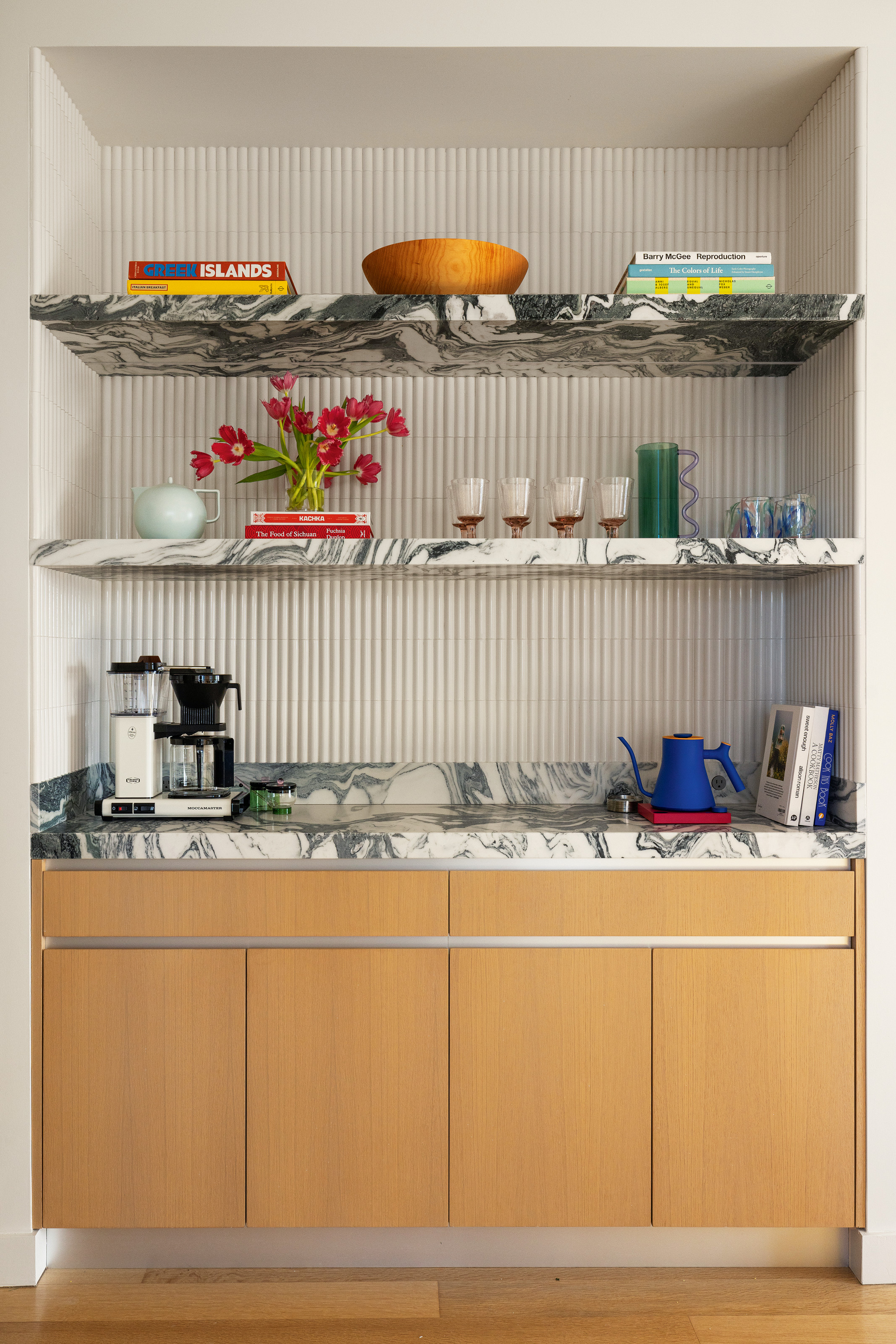 Corner of kitchen with floating oak cupboards, grey and white marble countertop and shelving and white fluted tiled backsplash