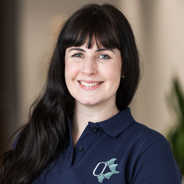 A professional headshot of a smiling woman with long dark hair and a fringe, wearing a navy branded polo shirt.