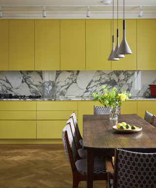 Yellow painted kitchen cabinets with marble backsplash, wooden flooring and a dark wood dining table and chair set.