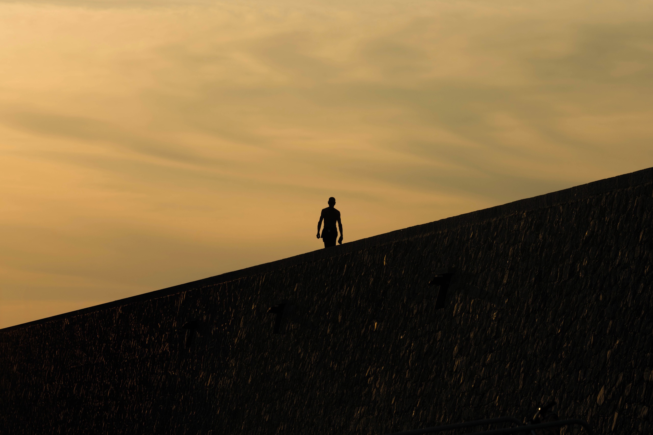 A lone silhouetted figure walks along the ridge of a sloping stone rooftop against a hazy orange sunset sky.