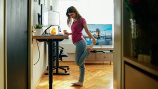 A woman stretches her left leg while standing at a standing desk. 