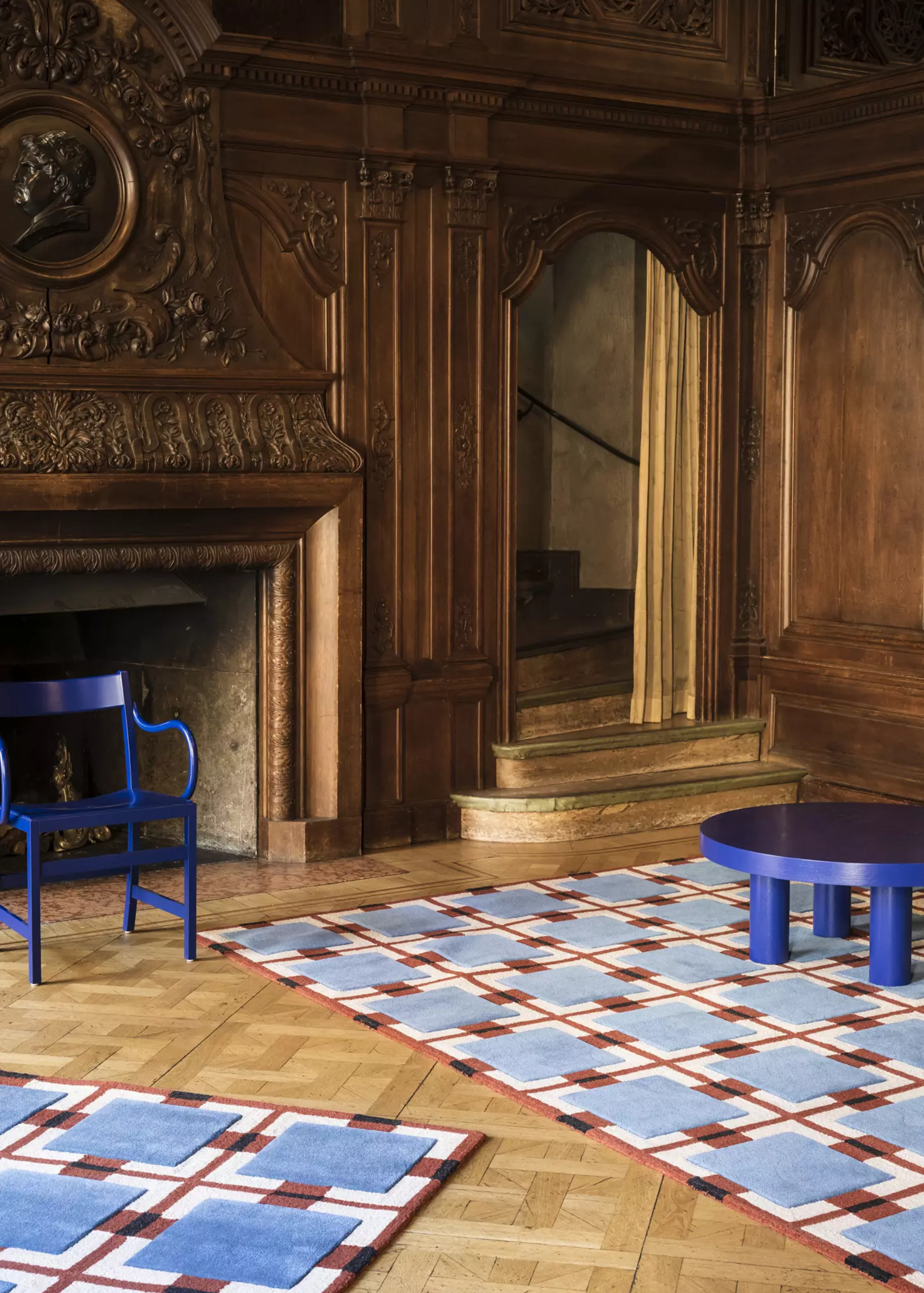 A living room with traditional woodwork on the walls and an ornate fireplace by cobalt blue armchair and a round coffee table on patterned rugs