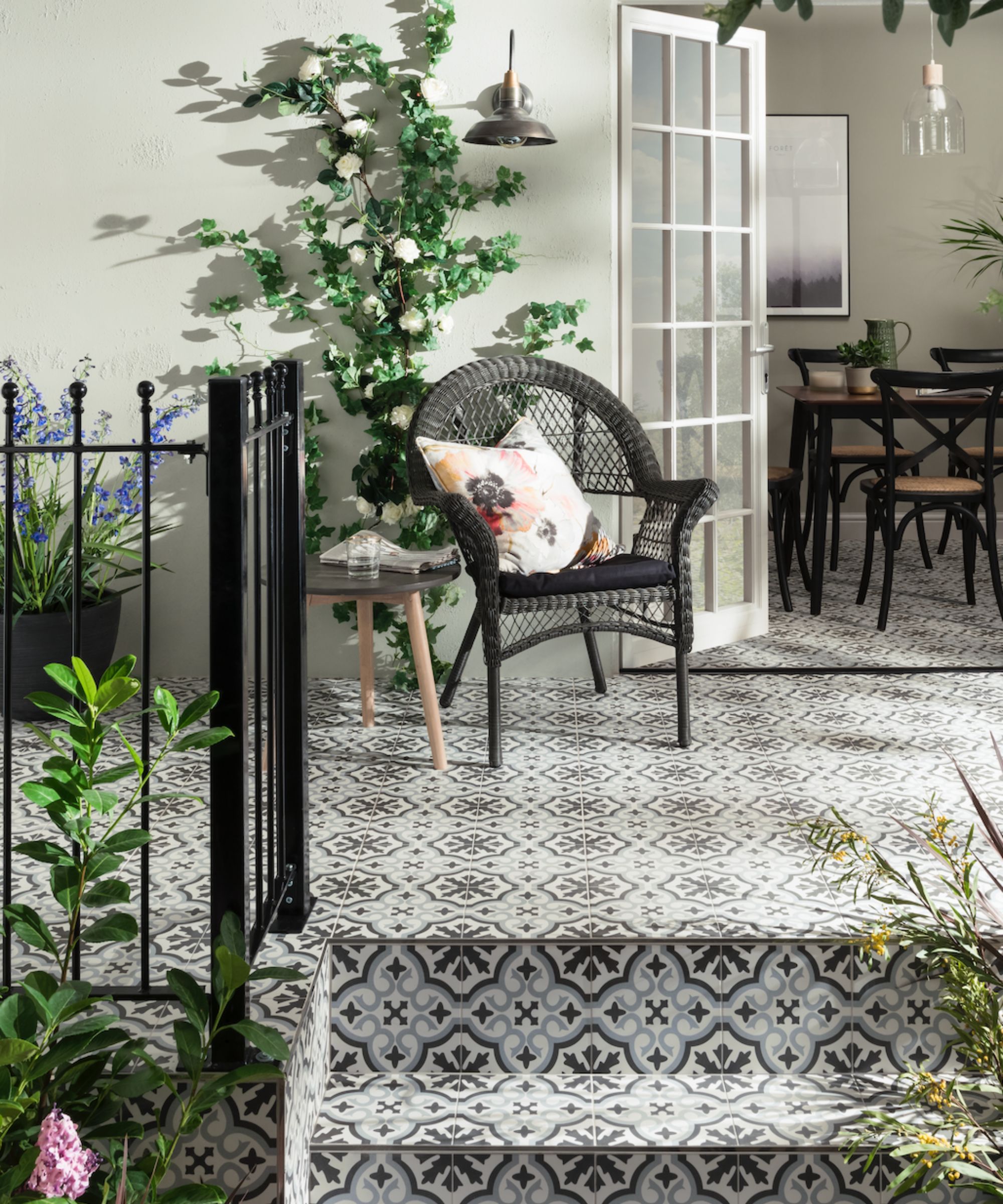 A patio with monochrome patterned tiles, a black rattan chair with floral cushion, climbing ivy, and French doors opening to a dining room.