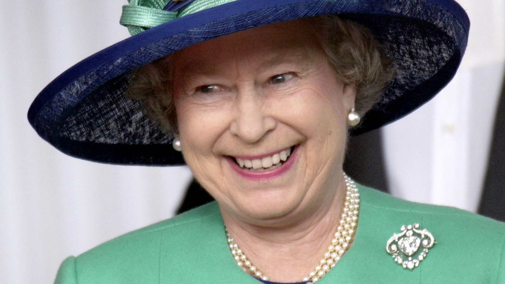 Queen Elizabeth wears a green jacket with a green and navy hat and laughs
