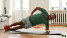 A man smiles and performs a side plank on an exercise mat at home. He is on his side, feet stacked one on top of the other, left forearm on the ground and body elevated. 