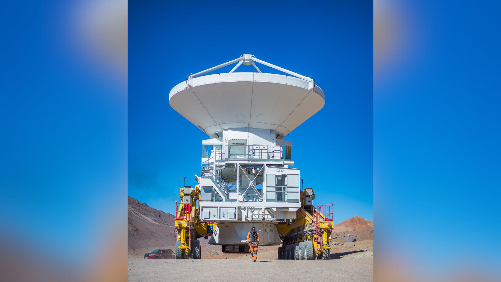 A large white radio dish sits on a transporter with giant wheels that rolls behind a man wearing an orange jumpsuit all in the hot sandy desert