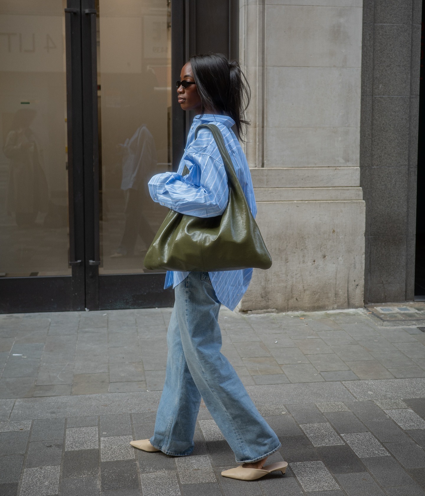 Influencer @_livmadeline stands outside in London wearing blue baggy jeans with beige low-heel mules, an oversized shirt and a large olive green handbag.