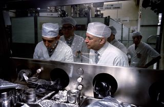 Dr. Robert R. Gilruth, on the right gets ready to inspect lunar samples along with several other scientists, all wearing protective white clothing.