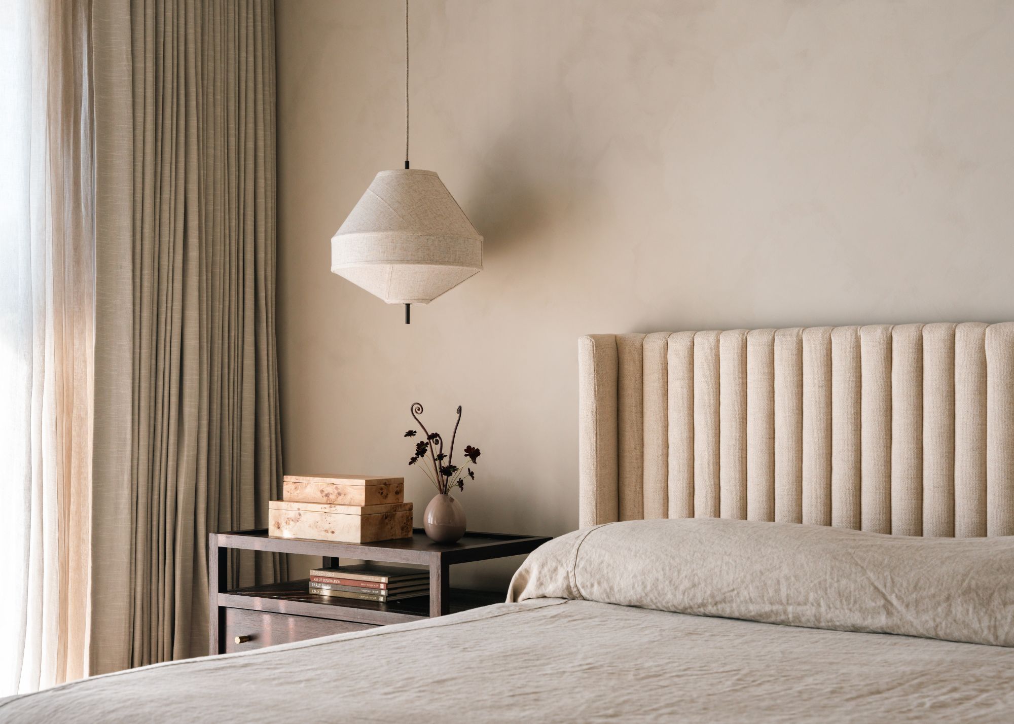 A minimalist Scandi bedroom with a linen pendant lamp, a ribbed headboard, a greige duvet, a metal nightstand, burl wood boxes, and a vase