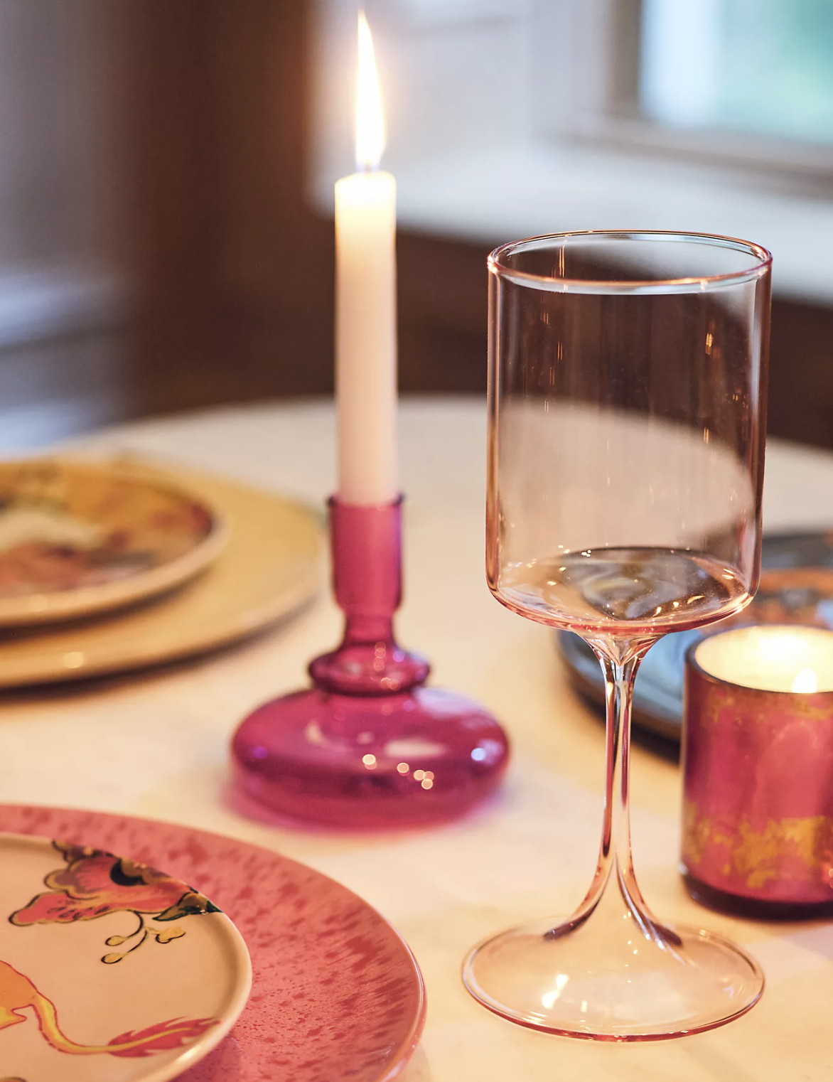 Meja makan dengan lilin magenta, tempat lilin, piring, dan kaca berwarna merah muda