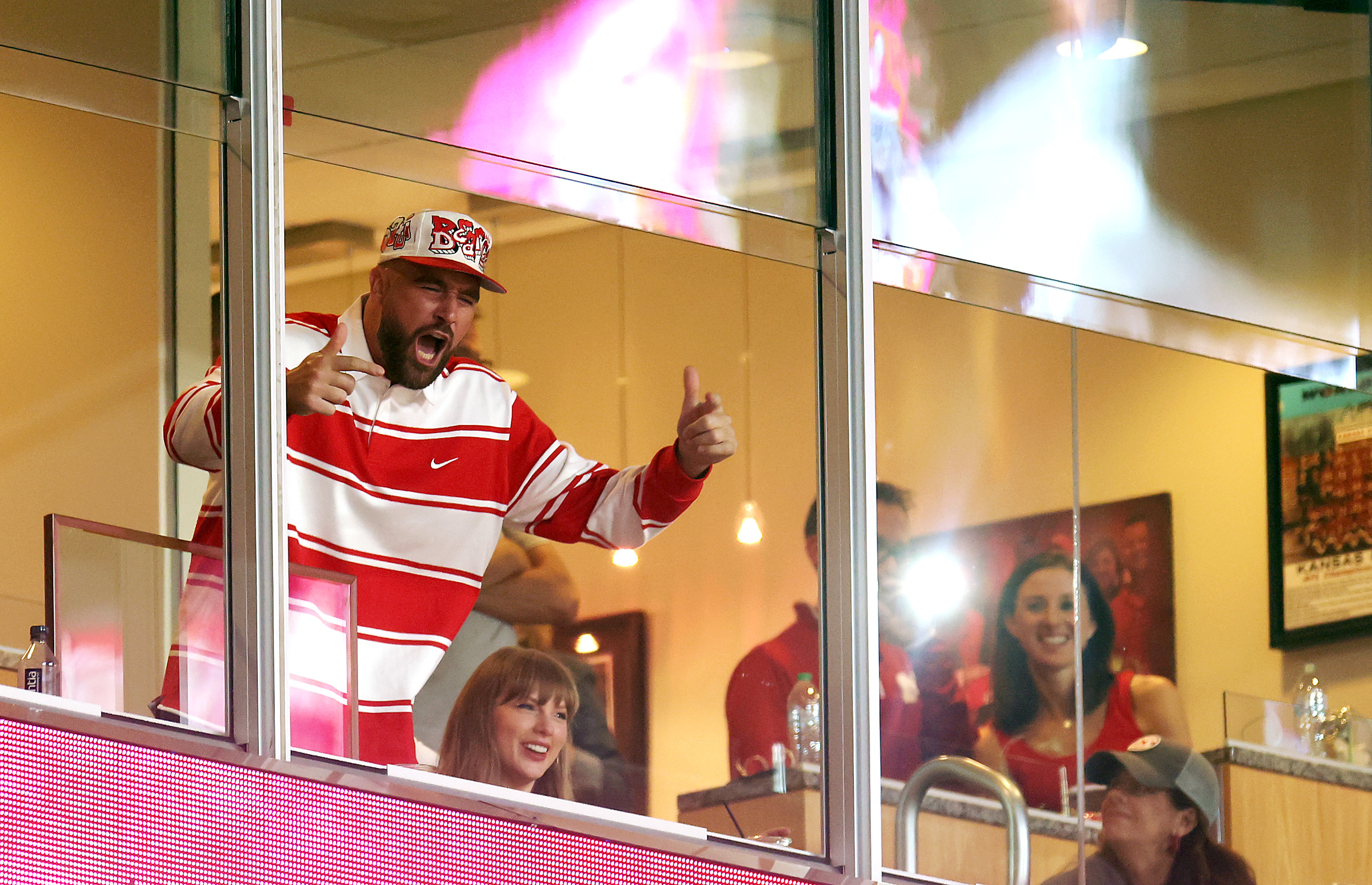 Travis Kelce wearing a red and white shirt and cheering while Taylor Swift sits next to him in a stadium box