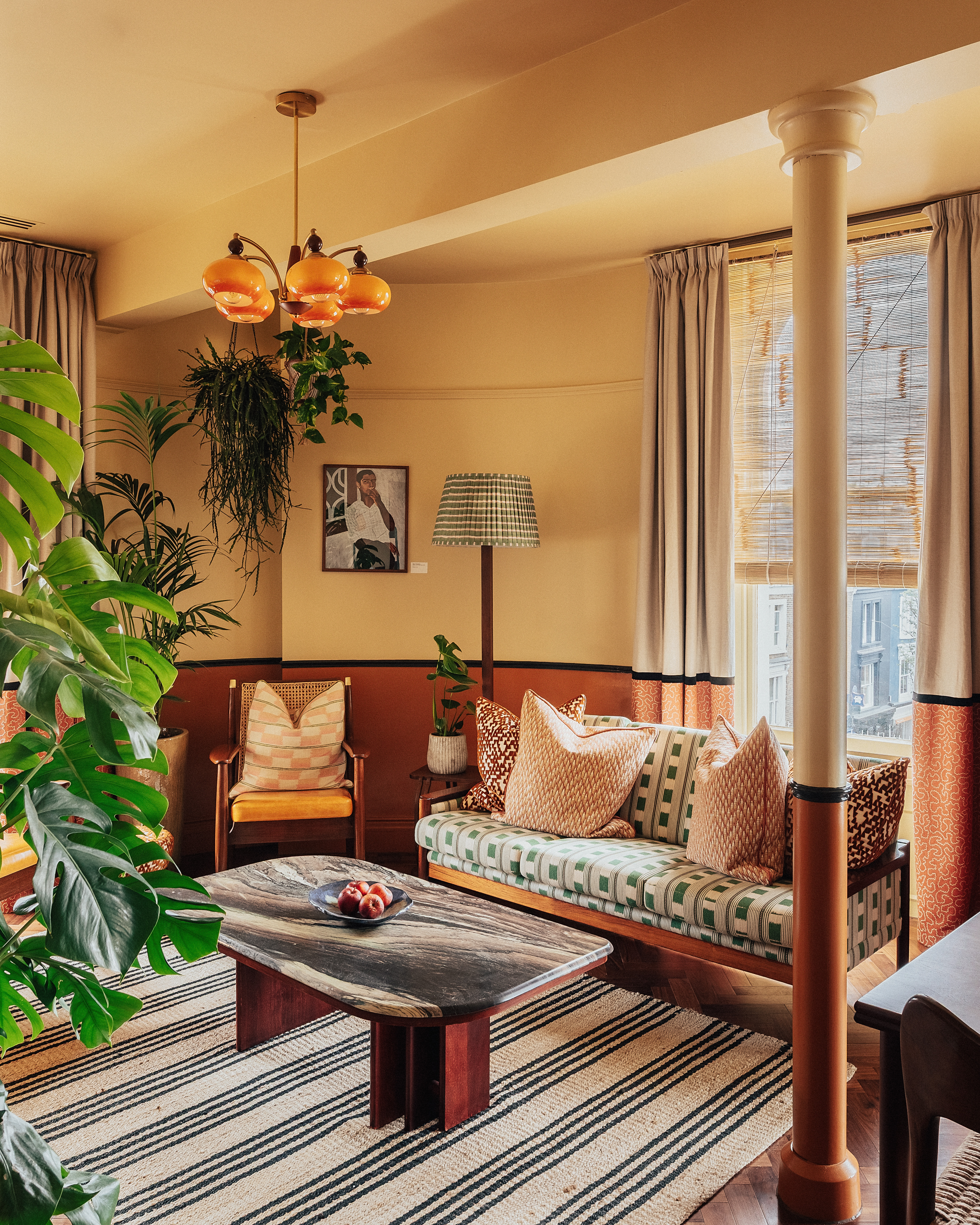 A layered, transitional hotel suite featuring a series of figurative, colorful artworks on its walls, patterned furniture, including rugs, armchairs, and sofas, split-color pillars, and a buttery, tile blue, and green palette.