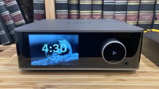 WiiM Ultra music streamer on wooden hi-fi rack in front of bookcase showing time on screen