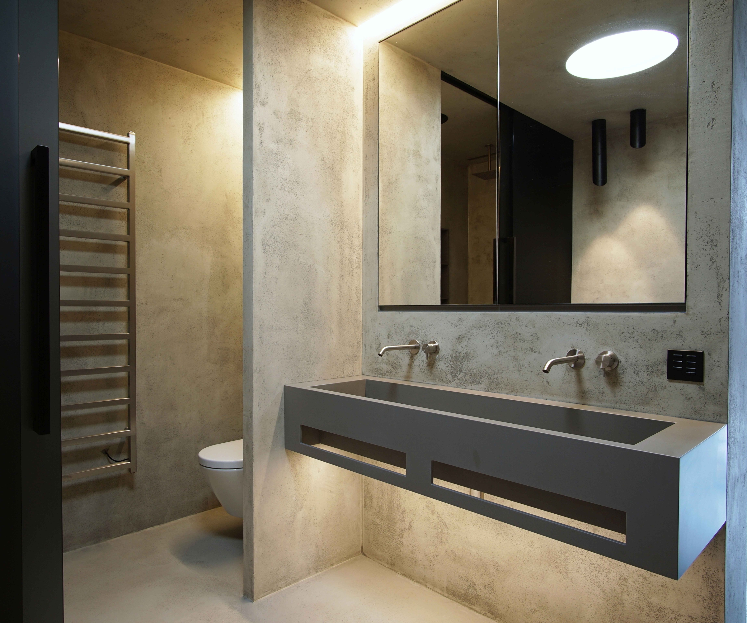 Concrete-textured bathroom with a floating grey trough sink, backlit mirror, and wall-mounted toilet.