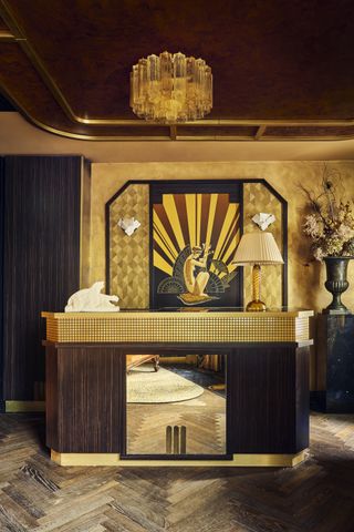 an art deco hotel reception with gold and timber desk with mirrored surface, herringbone floors, gold fabric walls, geometric shapes, a timber ceiling and glass pendant light